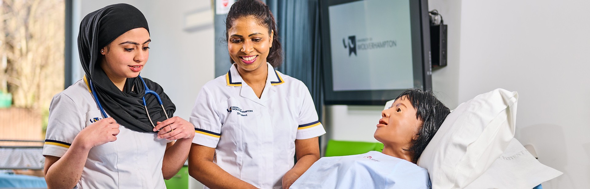 Two nursing students in uniform, standing over a demonstration dummy in a practical lab