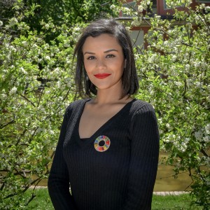 Nathalie Flores standing between two tall hedges, wearing a black blazer with a multicoloured brooch