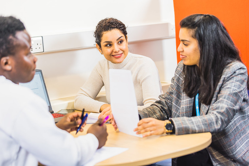 Three people sat together at a table, looking across at each other and conversing.