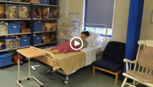 A dummy patient lying on bed with boxes next to him and a chair on the other side