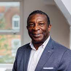 Professor Martin Levermore, MBE, photographed in front of a window with a cityscape behind him. He is wearing a smart shirt and suit. He is smiling jacket