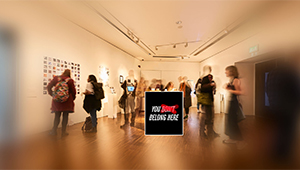 A group of people in an art gallery surrounding a sign where an exclusionary message has been corrected to read YOU BELONG HERE
