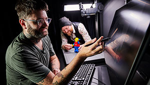 two males using the Animation stop motion facilities. IN the background, one person is positioning an oversized lego figure. Whilst the person in the foreground is recording this movement using a computer terminal.