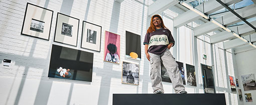 A photograph of Photography graduate Chandni Raithatha standing on top of a black plinth. She is pictured in front of her artwork - a series of framed photographic images a mixture of portraits and images of everyday objects