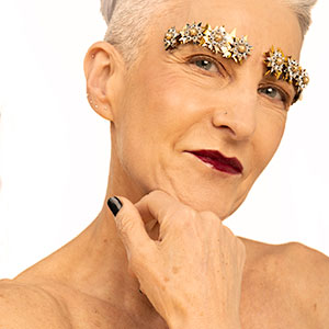 Photograph of a person with black fingernails and bejewelled eyebrows facing the camera with their hand placed under their chin.