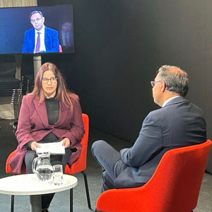 Photograph of an interview in progress, two people sitting in red chairs around a short table with the interviewee pictured on a screen behind the interviewer