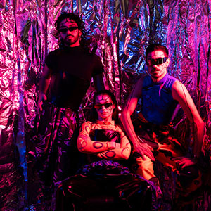 Photograph of three models in different outfits with matching shades and lined face paint, posed together under blue and purple lights against a foil background.