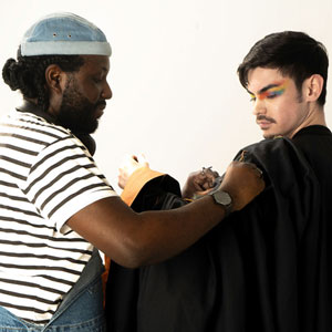 Colour photograph of a student with multicoloured eyeshadow being dressed in graduation robes.