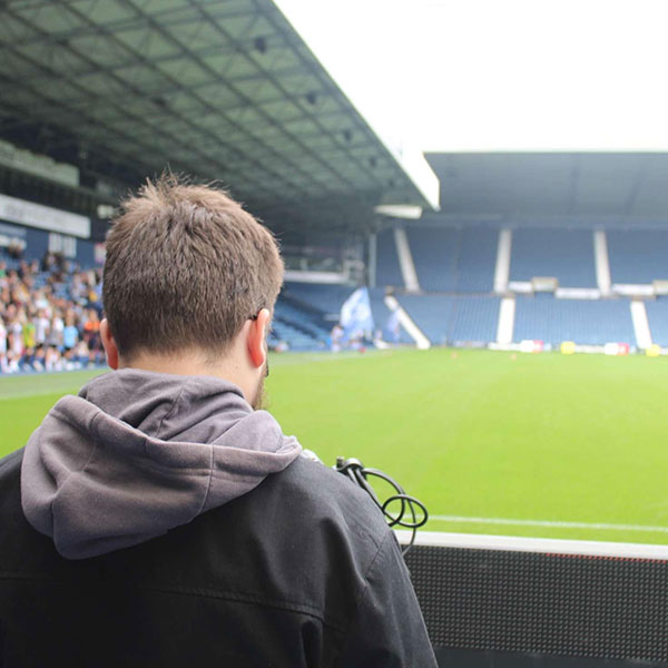Photograph by Callum Burgess of a person, seen from behind, looking out onto a football pitch visible over their shoulder