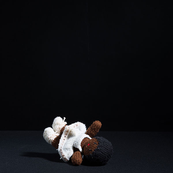 Photograph of a knitted doll wearing a ballet outfit, left on its back in a black room.
