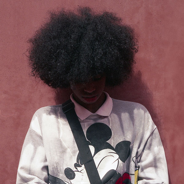 Photograph of a young person standing against a red wall and looking down at the ground.