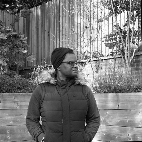 Black and white photograph of a person in a coat standing in a fenced garden.