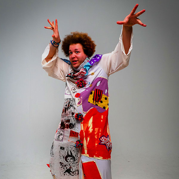 Photograph of a person in a white studio with their arms outstretched at their sides, wearing a patchwork outfit of various artworks.