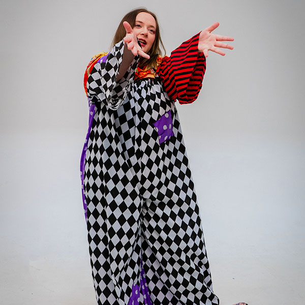 Photograph of a person in a white studio reaching forward, wearing a baggy checkerboard-pattern outfit with red stripe and purple polka-dot patches sewn sporadically into it.