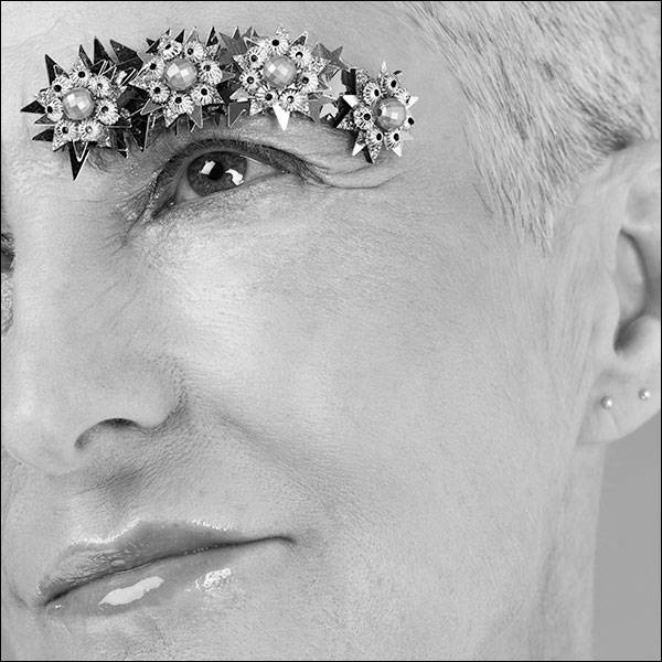 Black and white photograph of a person whose eyebrows are decorated with sequins and jewellery.