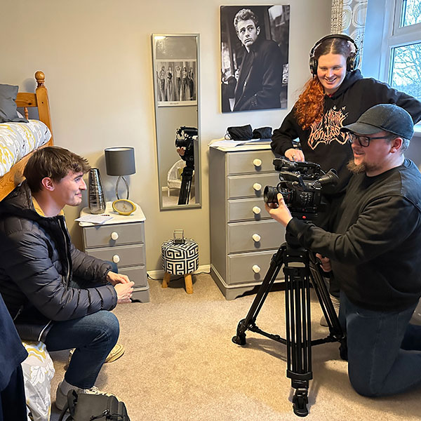 Three people with a camera on a tripod in a bedroom, two stood behind the camera filming one sat on a stool in front, one wearing headphones, next to a mirror and a poster of James Dean above a chest of drawers.