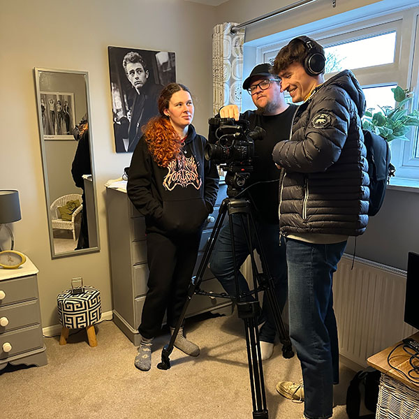 Three people with a camera on a tripod stood together in a bedroom, one wearing headphones, next to a mirror and a poster of James Dean above a chest of drawers.