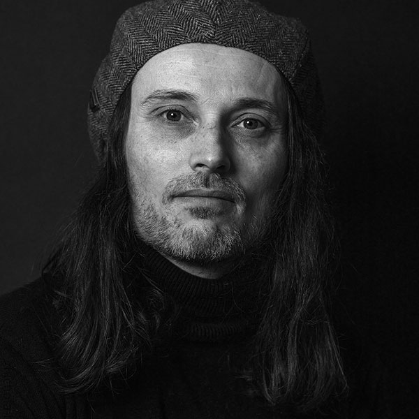 Black and white portrait photographed of a long haired person wearing a hat.