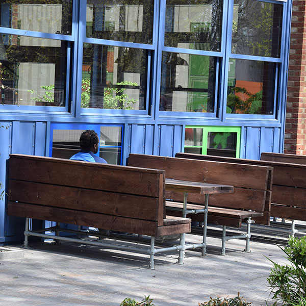 Photograph of a man sitting alone in an outdoor seating area next to the large windows of a building, wearing a shirt in matching blue tones to the window frames.