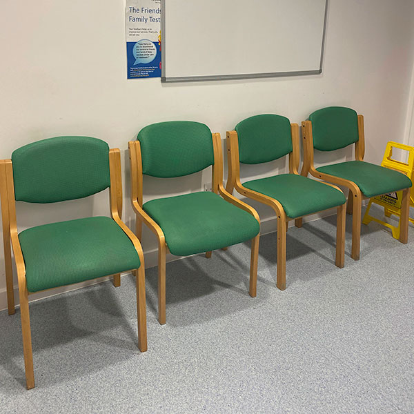 A row of four green chairs in a white waiting room, a small informative poster and whiteboard pinned to a wall.