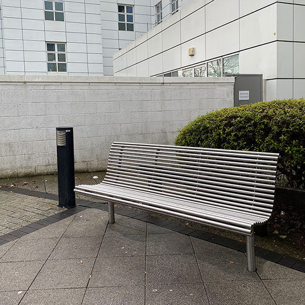 A silver metal bench on a footpath outside a large grey building.