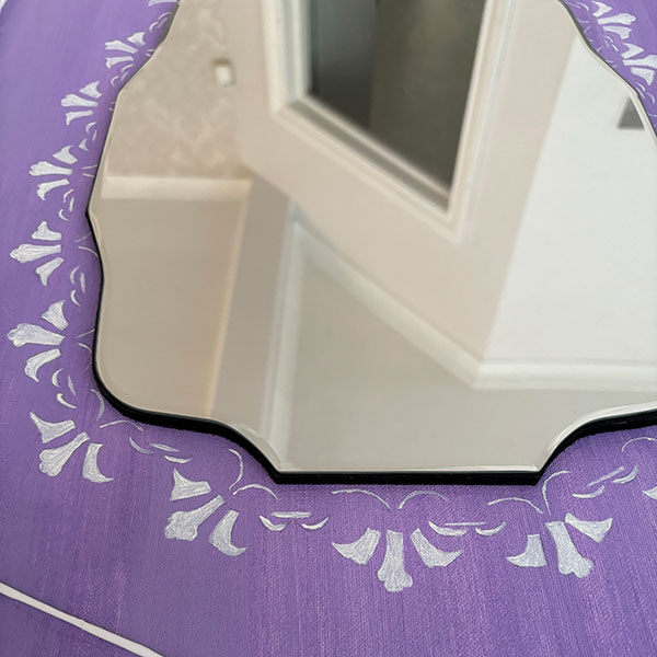 A mirror placed on a purple surface with ornate white patterns etched around its border.