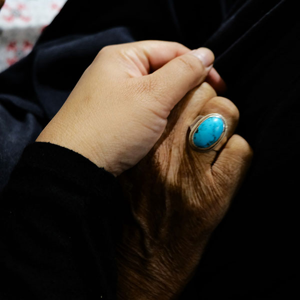 Two hands, one of a younger person clutching another of an older person, the older hand wearing a ring with a large blue stone.