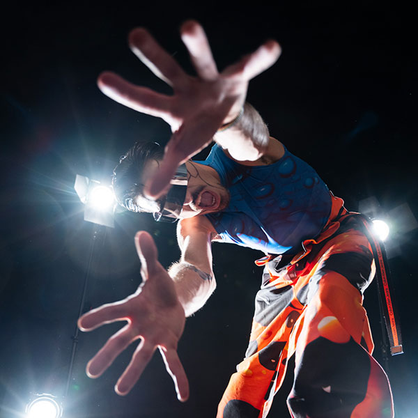 A person in black and orange patterned trousers, a blue vest with circular bubble patterning, visor shades and face- and body paint, arms outstretched towards the camera beneath them.