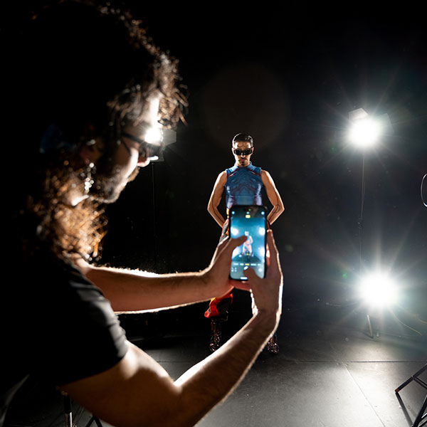 A person in a blue vest and visor shades posed with their arms behind their back in a circle of lights, being photographed by another model in the foreground using their phone.