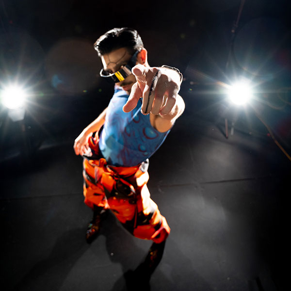A person in black and orange patterned trousers, a blue vest with circular bubble patterning, visor shades and face- and body paint, pointing towards the camera while being lit from behind.