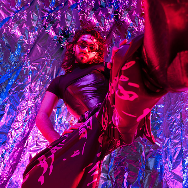 Photograph from below of a model in a small black top and patterned trousers, wearing shades and with their face painted into symmetrical segments.