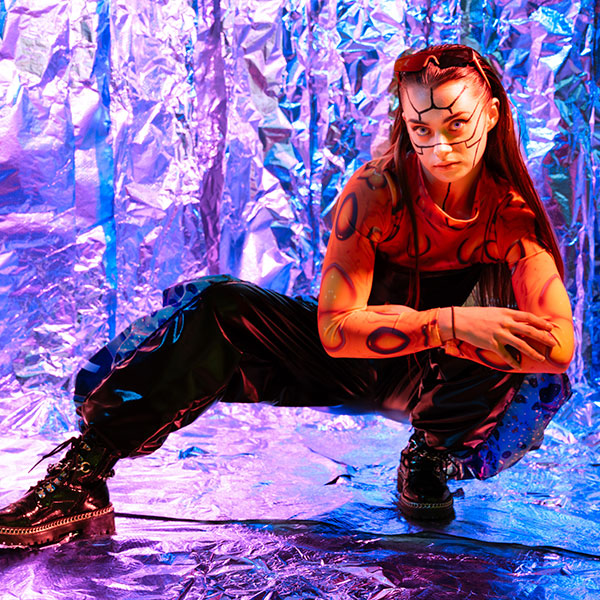 Photograph of a person in chunky black boots, baggy waist-high trousers and a top decorated with circle patterns, holding a low pose with their arms crossed, their face painted with symmetrical lines as if segmented, a pair of orange sunglasses on their head, photographed against a foil backdrop under blue and purple lights