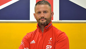 Paralympic Coach Ian Johns, photographed against a multicoloured background. Ian is is stood arms folded looking serious.
