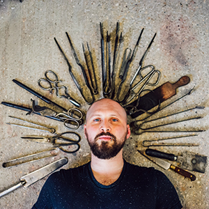 Portrait of Glass graduate, Elliot Walker he is pictured with a variety of glass sculpting tools in an arch above his head
