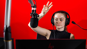 Female Multimedia Journalism student operating the control deck in the radio studio. They are wearing headphones and have their right hand raised, signalling to begin recording. They are photographed against a red background