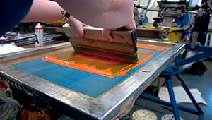Close up of someone screenprinting in the print studios. In the image you can see a squege drawing orange ink forwards on a blue screen surrounded by a metal frame.