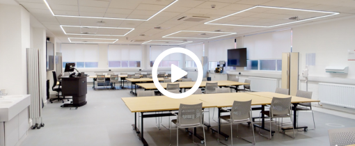 A lecture room with student desks, a lecturer desk and an interactive whiteboard, a play button icon identifying this as a video link