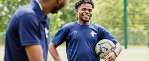 Two students in blue sporting clothes standing outside, one holding a football under their arm.