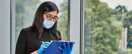 A student wearing glasses, protective gloves and a face mask, inspecting a document on a clipboard.