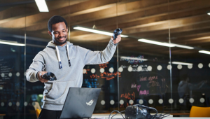 A student wearing a grey hoodie holding two motion control remotes while using a laptop.