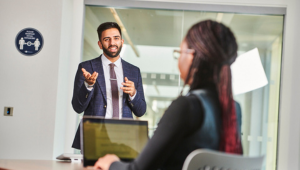 A student wearing a suit and tie, giving a presentation to a seated person at a laptop in an office room.