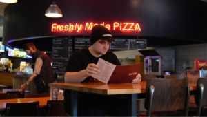 A student examining the menu in the Light cinema pizza restaurant, a neon sign in the background reading FRESHLY MADE PIZZA