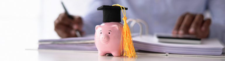 Student sat at desk reviewing documents in a binder. A pink piggybank stands in the foreground wearing a graduation cap.