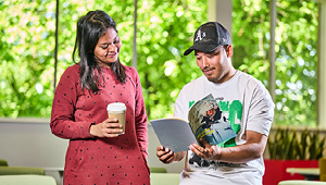 Two students stood looking over a guidebook, one has a coffee cup in hand