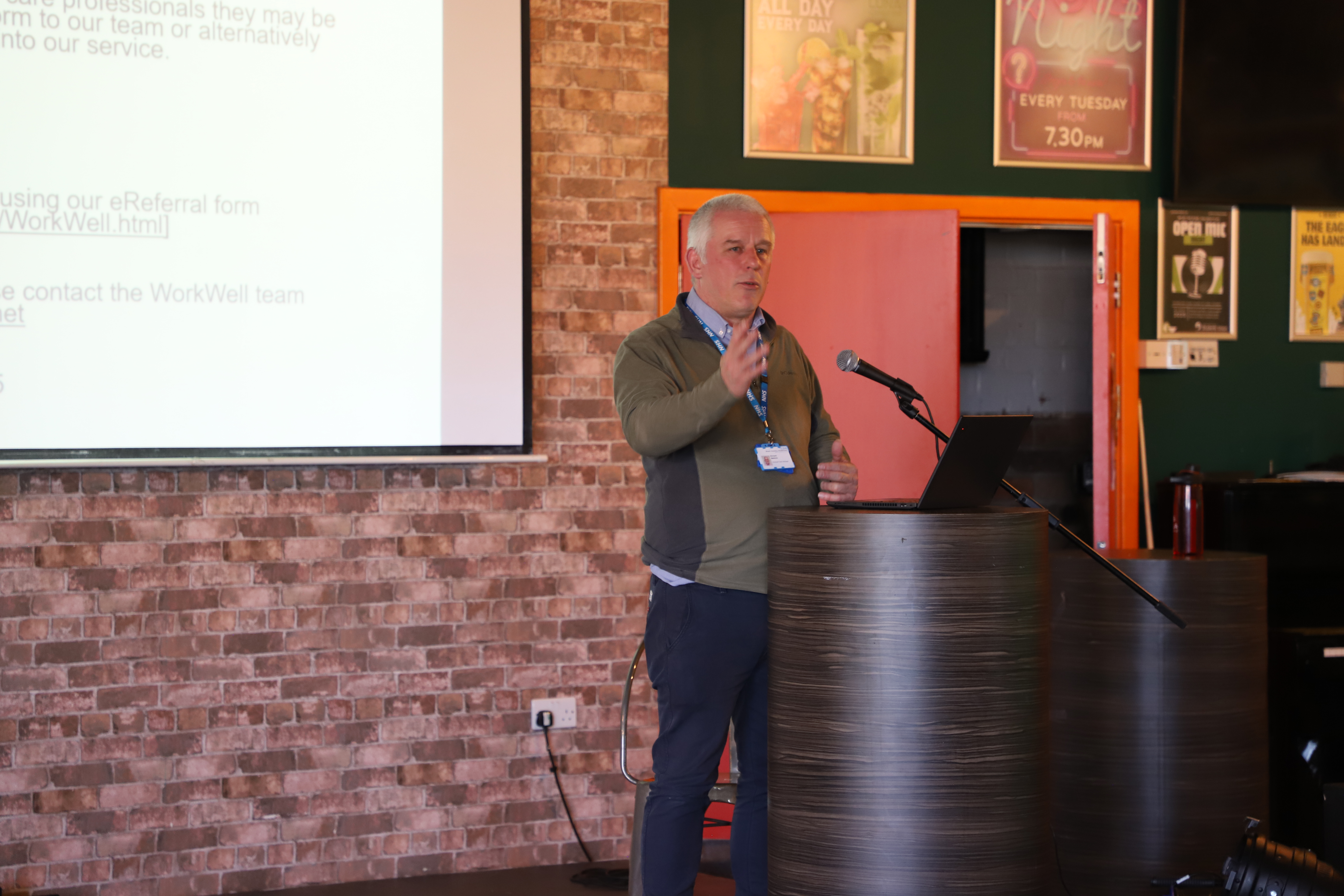 Work Well representative, male wearing a green jumper and blue trousers, standing on a stage delivering a talk