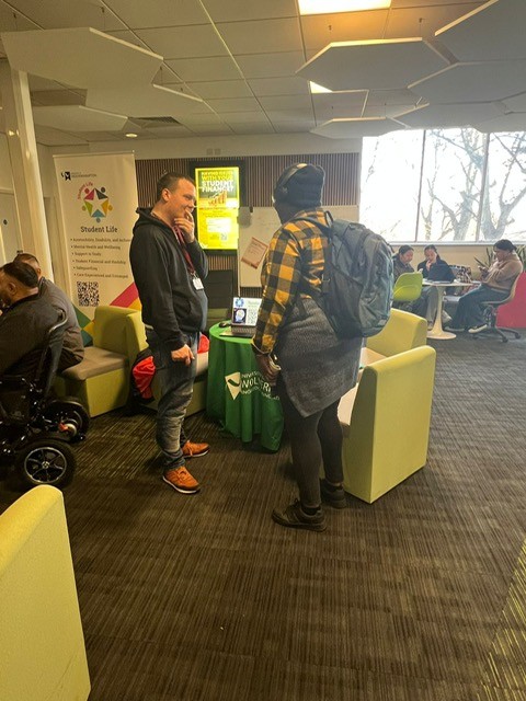 Student representative for Tourette's speaking to a event visitor, both make, standing and talking in a open space