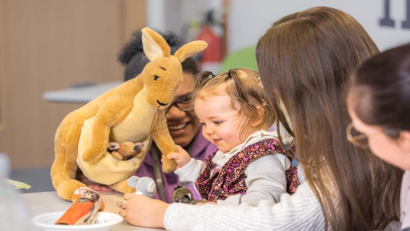 A child at a table supported by teaching staff and holding a plush toy kangaroo