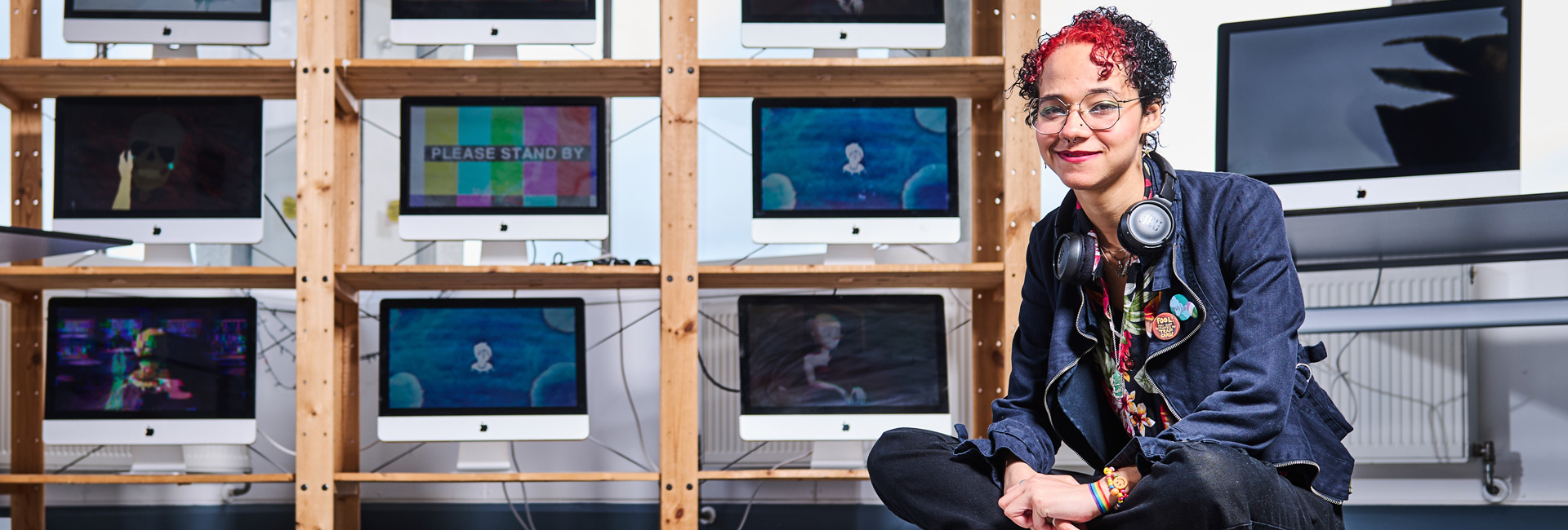 A photograph of a student sitting crossed legged on the floor, they have mixed black and red hair and are wearing glasses. There is a pair of headphones around their neck and behind them are nine monitors displaying different images.