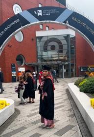 Graduate Aishah, standing outside university building in graduate gown and hat and smiling at camera