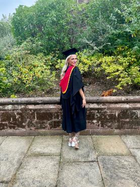 Graduate Kizzy standing outside with leaves in background wearing graduation cap and gown facing away from camera and looking back at camera and smiling